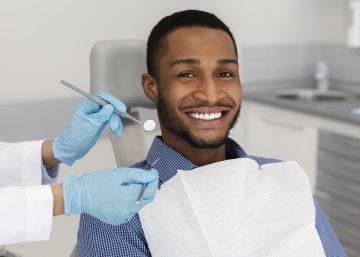 African american man smiling and sitting in dental chair while dentist holds tools